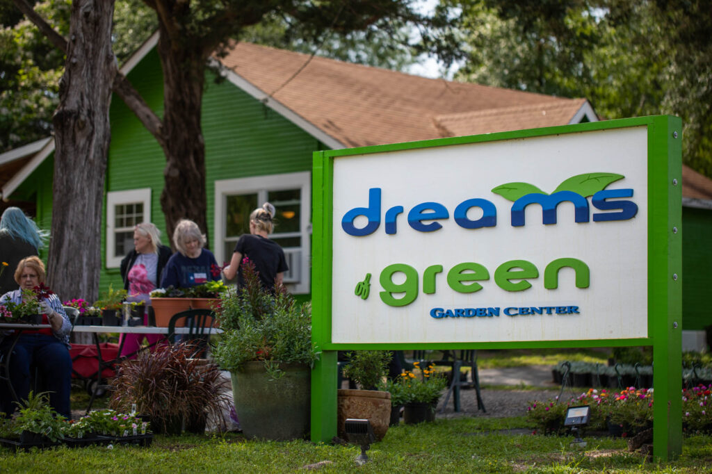 A bright green and white sign for "dreams of green GARDEN CENTER" stands in the foreground. In the background, people are gathered near a small, green building with a porch, surrounded by various plants and trees.