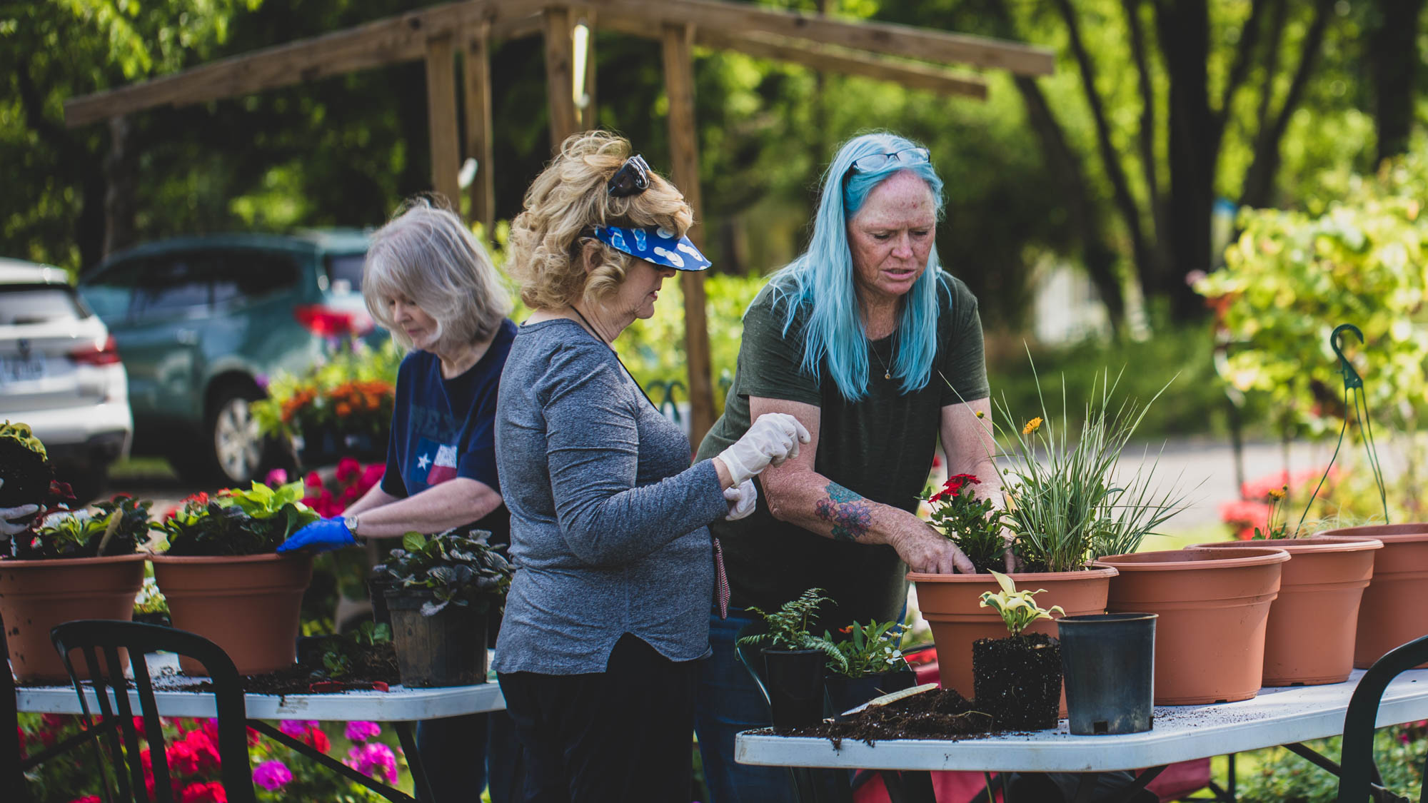 Three women are actively engaged in a hands-on gardening activity. The woman on the right with blue hair is focused on planting, the woman in the middle with a sun visor and white gloves is holding a small pot, and another woman on the left with blue gloves is tending to plants in a brown pot. They are all standing behind a long table with various plants and pots.