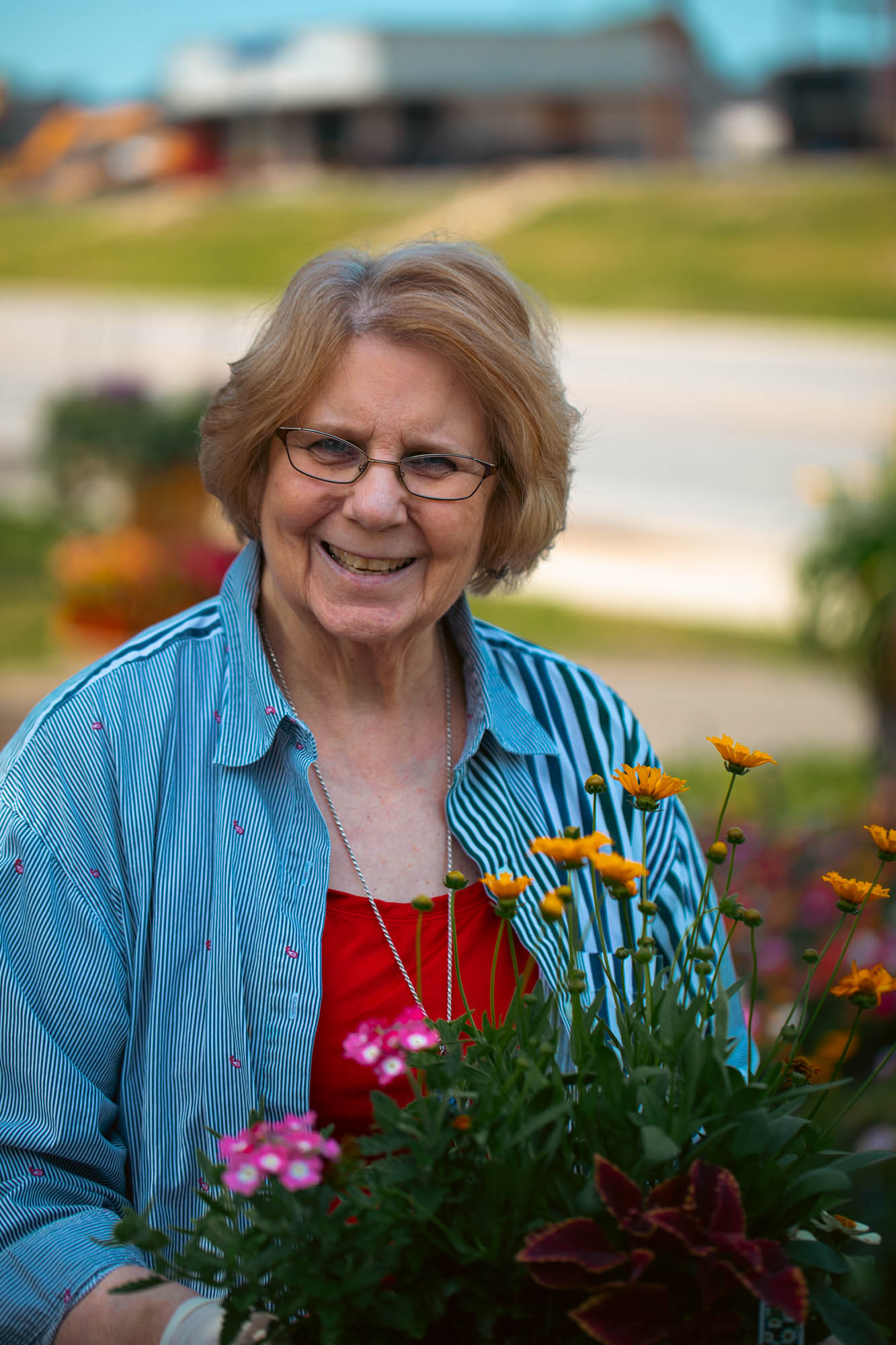 A smiling older woman with short blonde hair and glasses holds a container of bright orange and pink flowers. She is wearing a blue-and-white striped button-down shirt over a red top. The background is blurred, showing what appears to be a building.